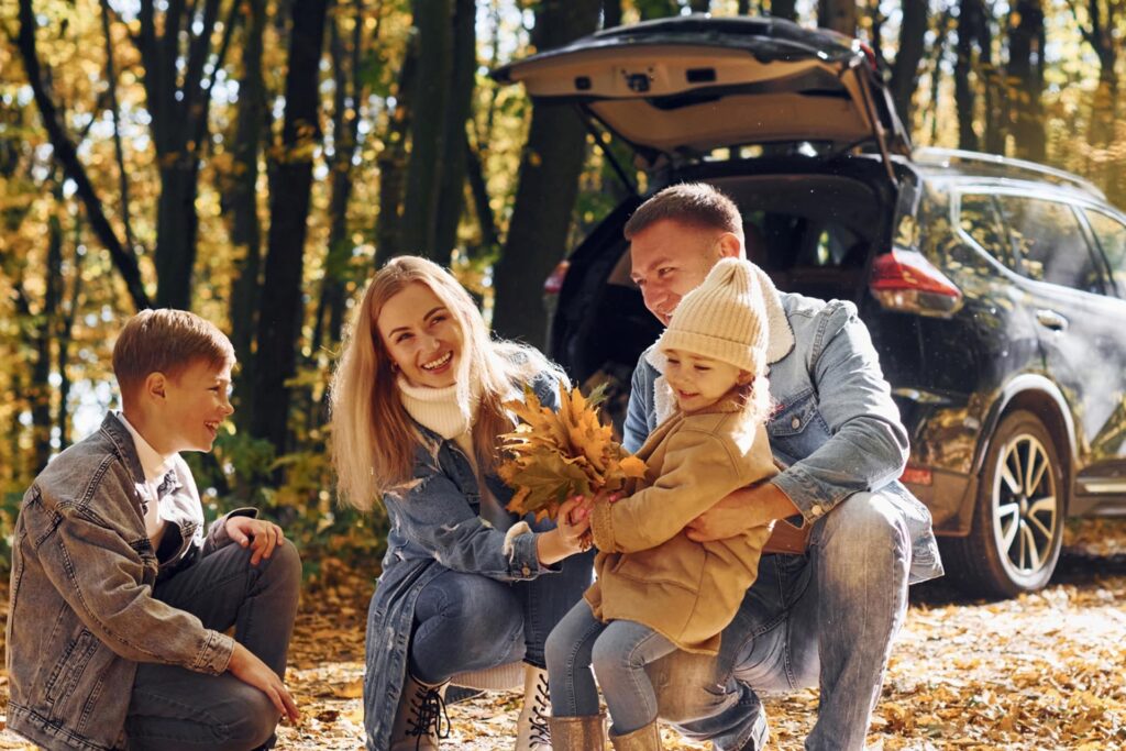 A family stands beside their car surrounded by colorful autumn leaves in a scenic fall landscape
