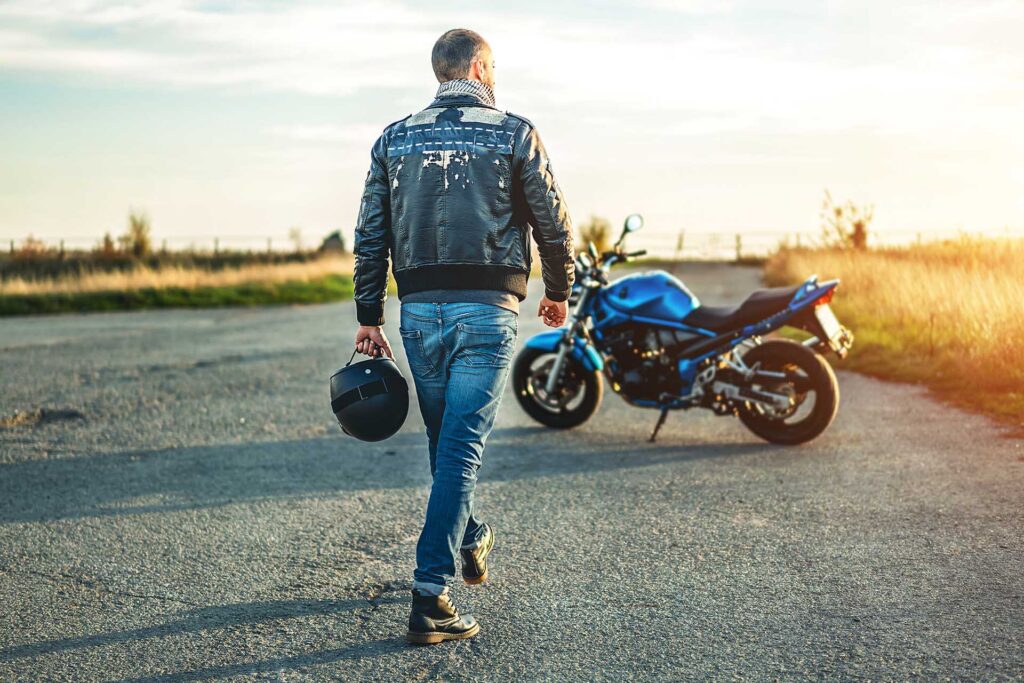 A man walks alongside a motorcycle on a road, surrounded by greenery and a clear blue sky