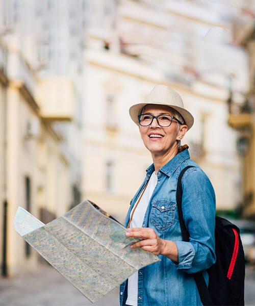 A smiling traveler holding a map outdoors, wearing a hat and glasses.