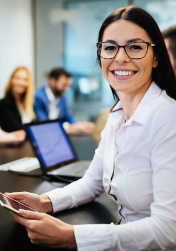 A smiling woman wearing glasses sits at a table, exuding a warm and friendly demeanor