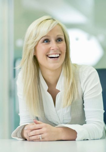 Smiling blonde woman in white shirt sitting indoors.